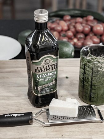 Rezept Foto Kräuterpesto. Das Foto zeigt viele Zutaten des Pestos. Im Vordergrund steht Parmesanreibe und ein Stück Grana Pandano und ein Messbecher voll mit frischen Kräutern.