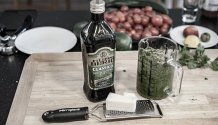 Rezept Foto Kräuterpesto. Das Foto zeigt viele Zutaten des Pestos. Im Vordergrund steht Parmesanreibe und ein Stück Grana Pandano und ein Messbecher voll mit frischen Kräutern.