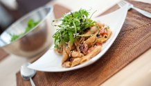 Rezept Foto vom italienischen Nudelsalat. Das Foto zeigt eine Nahaufnahme von einem gewölbtem Teller mit reichlich Nudelsalat darauf. Der Nudelsalat ist mit frischem Ruccola und Balsamico Creme garniert worden.