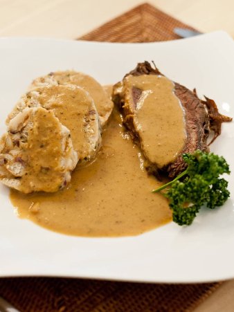 Eine Draufsicht auf den Sauerbraten, angerichtet auf einem großen weißen, geschwungenen Teller mit einem Semmelknödel und viel rahmiger Soße.