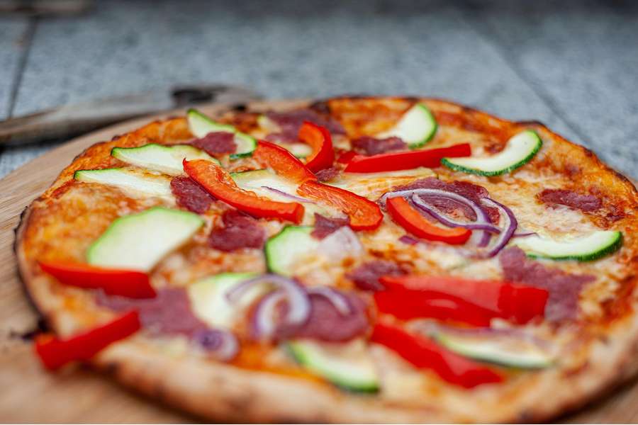 Rezept Foto Holzofen Pizza Komplettanleitung. Detailaufnahme. Fertig gebackene Pizza mit Salami und frischem Gartengemüse auf einem Holzbrett angerichtet.