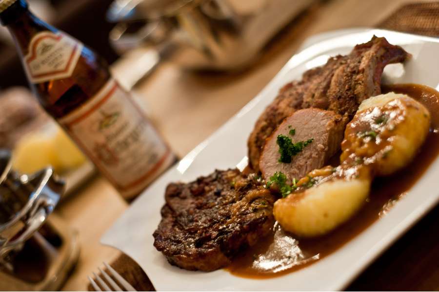 Rezept Foto Bayerischer Schweinebraten in der Kümmelbiersoße. Das fertige Gericht angerichtet auf einem geschwungenen Teller. Zwei Stücke Schweinebraten und einen Kartoffelknödel mit Kräutern bestreut aber in anderem Blickwinkel.