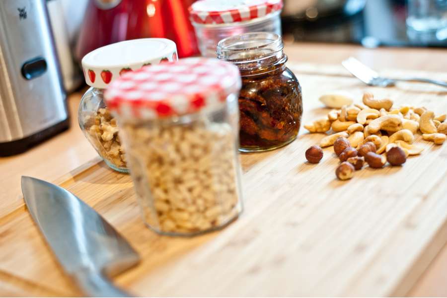 Rezept Foto Rotes Pesto Rosso. Nahaufnahme verschiedener Nüsse und Kerne. Walnüsse, Cashew Kerne, Haselnüsse und Erdnüsse. Die Gläser zum Abfüllen für das Pesto stehen auf einem Schneidebrett.